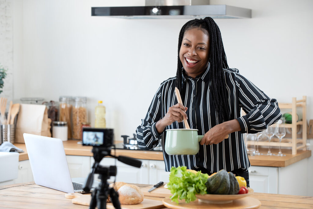 Happy african american woman vlogger broadcasting live video online while cooking food in kitchen at home