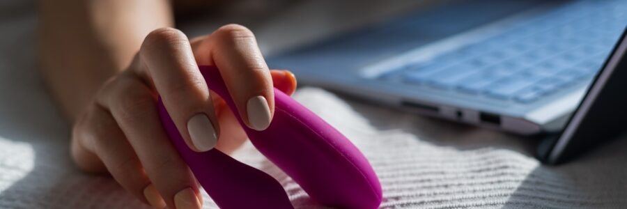 A woman lies in bed holding a clitoral vibrator and watching porn on a laptop. The girl has sex online.